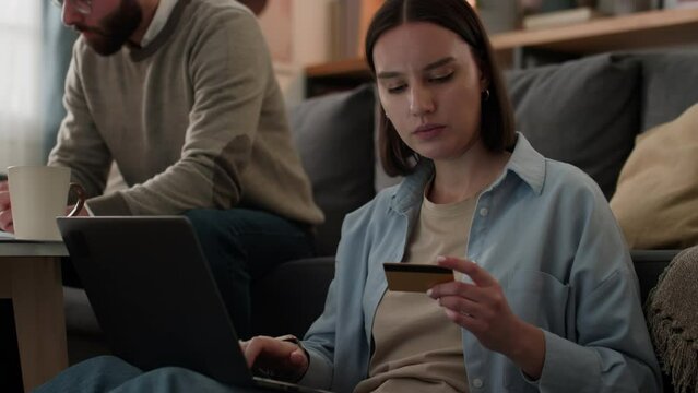 Medium close-up shot of young Caucasian woman sitting in living room with laptop, holding credit card, ordering from online store, chatting to husband, who is sorting utility bills, both laughing