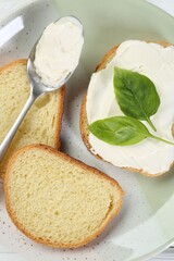 Pieces of bread with cream cheese and basil leaves on white wooden table, top view