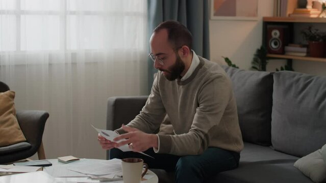Medium shot of young Middle Eastern man sitting on couch at home, dealing with household expenses, opening envelope, taking out new bill, examining it, clenching fist and swearing