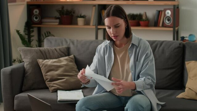 Medium shot of young Caucasian woman sitting on couch in living room, opening envelope with bank account statement or utility bill, examining it then cheering with joy and pumping fist