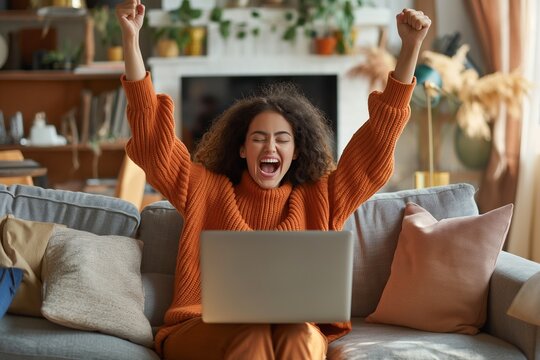 Excited woman sitting on a sofa, holding a laptop on her lap and making a "Yes" hand gesture. She screams with joy, feeling happy about getting her dream job, an email sales discount offer,