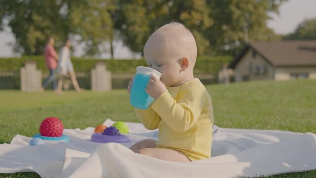 Fun Infant enjoy drink from blue sippy cup while seat on blanket, surround by toys. Cute baby boy drink water, sunny park setting emphasize playful, vibrant, and engage atmosphere of outdoor scene.