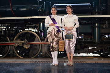 Two attractive women  in traditional Thai dress pose for a photo with a steam train at  the...