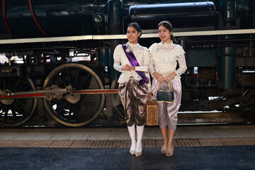Two attractive women  in traditional Thai dress pose for a photo with a steam train at  the platform of the retro train station