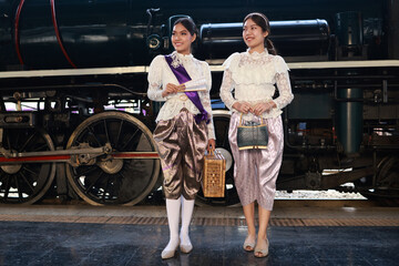 Two attractive women  in traditional Thai dress pose for a photo with a steam train at  the platform of the retro train station