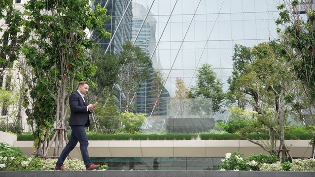 Side view of caucasian businessman looking at smart phone and walking at modern building in green eco city. Skilled leader going to workplace while checking report by using phone. Lifestyle. Urbane.