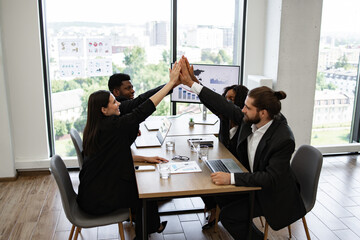 Business team giving high five around a meeting table in a modern office with city view. Celebrating a successful project.