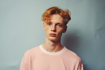A young man with red hair and a pink shirt is standing in front of a blue wall