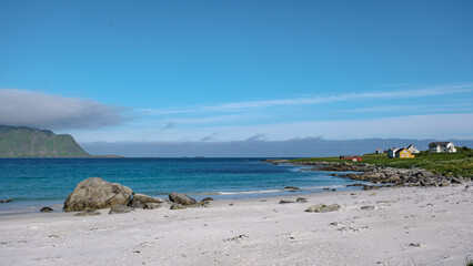 Stunning white sand beach in Norway with clear blue water, green hill, colorful houses, and blue sky with white clouds. Ramberg Beach Lofoten Norway