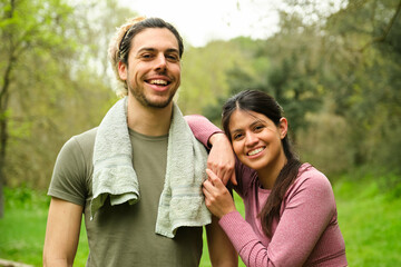 Obraz premium A man and a woman runners are smiling and looking at camera in a park. The man is wearing a green shirt and a towel, while the woman is wearing a pink shirt. Scene is happy and friendly