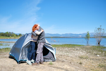 A man and woman hug in front of a tent by a lake. The scene is peaceful and serene