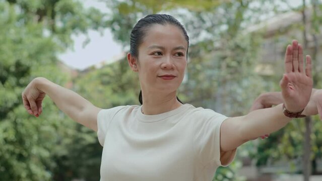 Medium shot of active middle-aged Asian woman performing tai chi arm moves in local city park