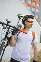 Portrait of guy wearing a helmet and sunglasses with bicycle against grey wall.