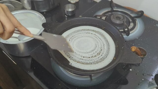 hand of a woman preparing authentic dosa in the kitchen in a frying pan under a stove using a ladle for breakfast