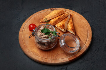 Homemade pate or mousse made from turkey or chicken liver in a glass jar. Baguette slices. Cherry tomatoes on a wooden round board.