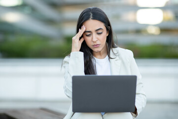 A young woman in a white blazer sits outdoors, using a laptop computer. Her expression is serious and focused, and she appears to be concentrating on the screen.