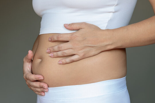 Stomach ache, bloating, flatulence, abdominal distension, colic, digestive disorders. Young woman in profile holding her stomach with hands on a grey background. Overeating, overweight, excess weight
