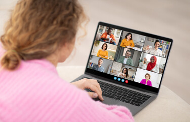 A woman is sitting at a desk and looking at a laptop screen. She is participating in a video conference call with eight other people. The people on the screen are in a grid layout