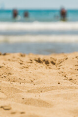 Beach sand with sea waves and bathers in the distant. Close-up of fine sand at the seashore. View as relaxing laying on towel sunbathing. Summer vacations. Vertical.