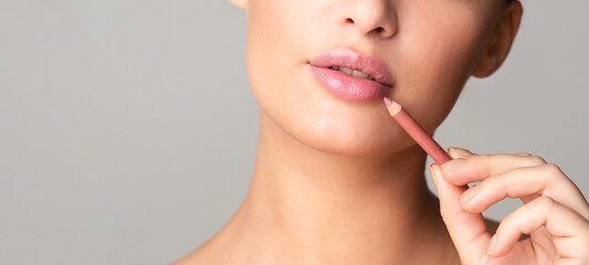 A close-up of a woman face as she carefully applies a pink lip liner to her lips, demonstrating a makeup technique. The background is plain and neutral, cropped, copy space