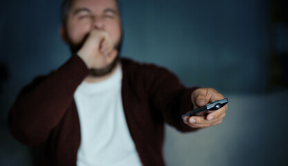 A bearded man wearing a white t-shirt and brown cardigan yawns while pointing a television remote, likely watching something late at night. The background is dark, implying a late hour.