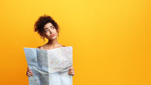 African American young woman with curly hair appears confused and uncertain as she holds an unfolded map in her hands, looking up and to the side as if seeking guidance, copy space