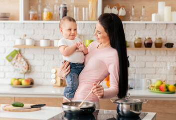 A woman is standing in a kitchen, holding a baby in her arms while cooking food on the stove. She is multitasking by caring for the child and preparing a meal simultaneously.