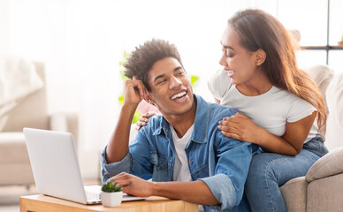 Black teen couple is relaxing at home. The guy is sitting at a table using a laptop, while the girl is sitting behind him, resting her arms on his shoulders. They are both smiling and laughing