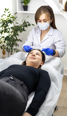 Cosmetician wearing nitrile gloves and a face mask, putting-on a hair cap on her client's head. The client is lying down on treatment table.