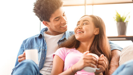 Loving teen couple enjoying coffee together in their living room. The guy is sitting on the couch with his arm around the girl, and they are both smiling and looking at each other