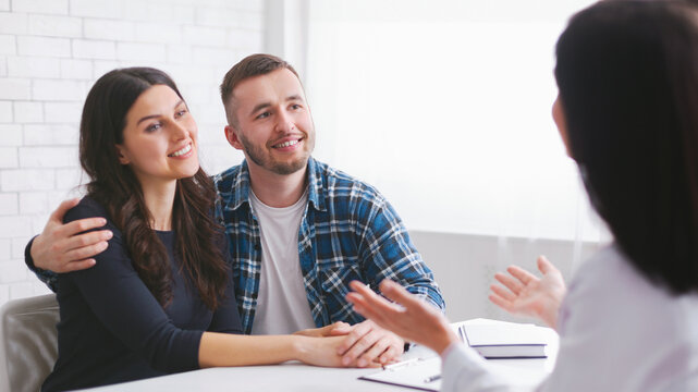 A young couple sits at a table listening attentively as a financial advisor explains something to them. The couple is smiling and appears to be engaged in the conversation