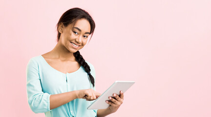 Fototapeta premium A young black girl is sitting against a pink background, focused as she uses a tablet computer. Her hands are tapping and swiping on the screen, with a look of concentration on her face.