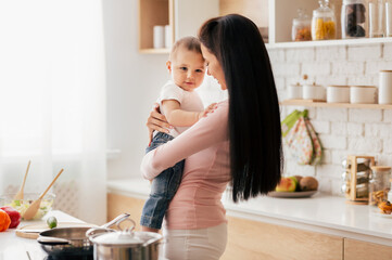 A woman standing in a kitchen, holding a small baby in her arms. The woman is dressed casually, and...