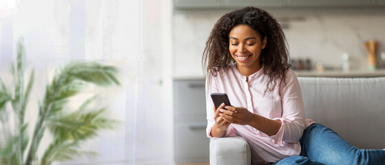 A happy black woman with curly hair is sitting on a light gray couch in a modern living room. She...