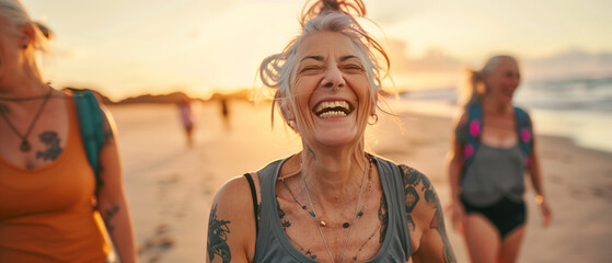 Laughing senior middle aged woman with gray hair and tattoos in sporty outfit walking on seaside beach with blurred aged friends in sunset. Aged people enjoy life. Active elderly people's lifestyle