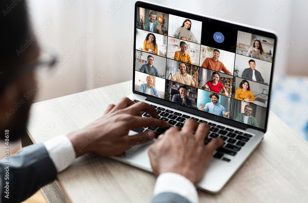 Aufkleber Black man sits at a table in front of an open laptop, actively participating in a video conference. The laptop screen displays a grid of various participants, each in their own frame