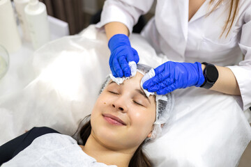 Naklejka premium Cosmetologist in nitrile gloves removing the remains of gel from the young woman's face using paper tissues, top view close-up.