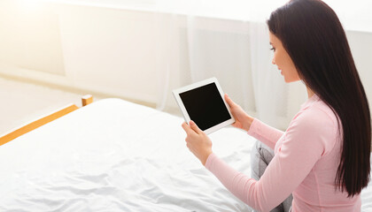 A woman is sitting on a bed, holding a tablet with black screen in her hands. She is looking at the screen intently, possibly reading or watching something, mockup copy space