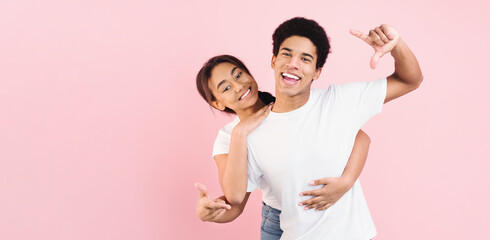 A joyous young African American couple is posing playfully against a solid pink background. The guy is making playful hand gestures, while the girl smiles warmly as she embraces him from behind