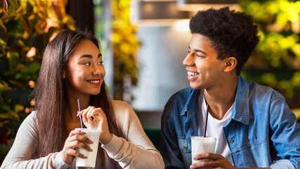African American guy and girl are sitting at a table in a cafe, each holding a glass of milkshakes. They are casually sipping their drinks in a relaxed atmosphere.