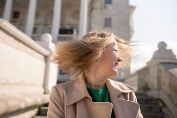 A woman with long hair is standing on a set of stairs. She is wearing a green shirt and a tan coat.