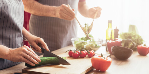 A couple is working together in the kitchen, preparing a fresh salad. The woman is using a knife to chop a cucumber on a wooden cutting board while the man is adding olive oil to a bowl of greens