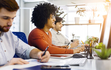 A diverse group of multiethnic men are seated around a desk, focused on a computer screen. They appear to be engaged in a collaborative work or discussion, with papers and notebooks scattered around.