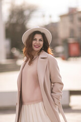 A woman wearing a white hat and a tan coat stands on a sidewalk. She has red hair and is smiling.