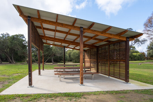 A wooden gazebo or pavilion with a roof, featuring public bench seats, serves as a covered picnic area in a park, offering ventilation and recreational rest facilities.