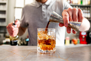 A close-up of a person pouring dark amber liquid into a glass with ice cubes on a bar counter.