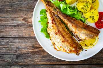 Fried sea bass served on lettuce with boiled potatoes and  lemon on white plate on wooden table
