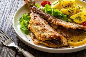 Fried sea bass served on lettuce with boiled potatoes and  lemon on white plate on wooden table
