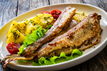 Fried sea bass served on lettuce with boiled potatoes and  lemon on white plate on wooden table
