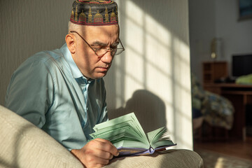 Lifestyle portrait of senior islamic man reading Quran at home. Natural aesthetic light